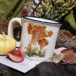 Puede incluir: Taza de cerámica blanca con borde y asa negros. La taza tiene un diseño moteado y una ilustración colorida de rebozuelos.