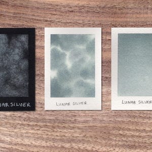 May include: Three watercolor paint swatches labeled "Lunar Silver" with different shades of gray. The swatches are on a wooden surface.