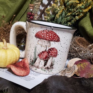 Puede incluir: Taza de cerámica blanca con borde y asa negros. La taza presenta un fondo blanco moteado con una ilustración en blanco y negro de tres setas rojas y blancas.