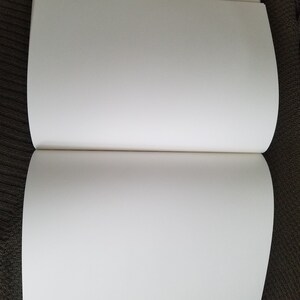Puede incluir: Un libro blanco de tapa dura, en blanco, con lomo y p&aacute;ginas. El libro est&aacute; abierto y acostado sobre una superficie marr&oacute;n.