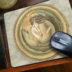 Peut inclure: Un tapis de souris rond avec une illustration aquarelle d'une sirène à la queue dorée, endormie dans un cadre circulaire avec une bordure dorée.