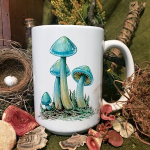 Puede incluir: Taza de cerámica blanca con una ilustración de setas azules y verdes. Las setas están representadas en un grupo sobre un fondo azul claro.