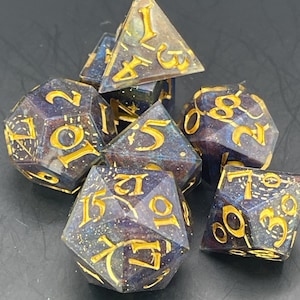 May include: A set of seven polyhedral dice featuring a deep blue and purple translucent resin base with embedded gold glitter particles. Each die has engraved numbers filled with a metallic gold paint, displaying various geometric shapes.