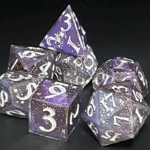 Puede incluir: Un conjunto de siete dados poliédricos con temática celestial. Los dados son una mezcla de resina morada y transparente, con números blancos y estrellas incrustadas. El conjunto incluye un D4, D6, D8, D10, D12, D20 y un dado de porcentaje.