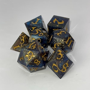 May include: A set of seven polyhedral dice with a celestial theme. The dice are a deep blue with gold numbering and inclusions, creating a galaxy effect. The dice are designed for tabletop role-playing games.