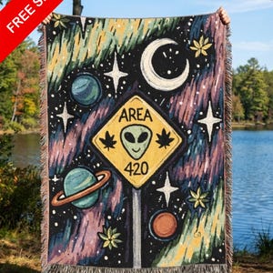 May include: A woven throw blanket with a space-themed design, including planets, stars, and a crescent moon on a dark backdrop. The blanket features a sign that reads "AREA 420" with an alien head. The blanket has a fringed edge.