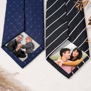 Könnte beinhalten: Zwei Krawatten mit personalisierten Fotoeinsätzen. Eine ist marineblau mit einem kleinen weißen Punktmuster, die andere ist schwarz mit weißen diagonalen Streifen. Jede Krawatte hat ein gerahmtes Foto.