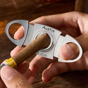 Pu&ograve; includere: Un tagliasigari argentato con il nome "Austin" e la data "13.2025" incisi. Il tagliasigari viene utilizzato per tagliare un sigaro marrone con una fascia gialla e bianca. Lo sfondo &egrave; una superficie di legno sfocata.