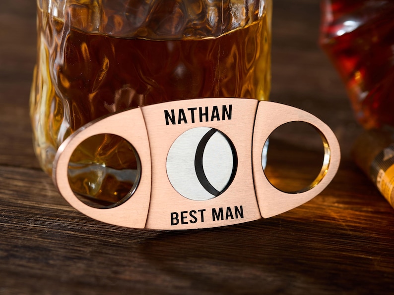 Pu&ograve; includere: Tagliasigari color rame con le scritte nere "NATHAN" e "BEST MAN". Presenta due aperture circolari e una lama centrale. Lo sfondo mostra un bicchiere con liquido ambrato.