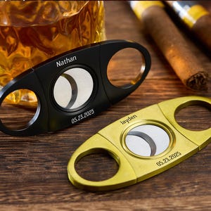 Pu&ograve; includere: Due taglia-sigari, uno nero e uno dorato, con testo personalizzato. Il taglia-sigari nero ha inciso "Nathan" e "05.23.2025". Il taglia-sigari dorato ha inciso "Jayden" e "05.23.2025". Sigari e un bicchiere di liquido ambrato sullo sfondo.
