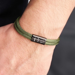 May include: Olive green braided bracelet with a black clasp. The clasp features a white cross, heartbeat, and heart symbol. The bracelet is worn on a wrist.