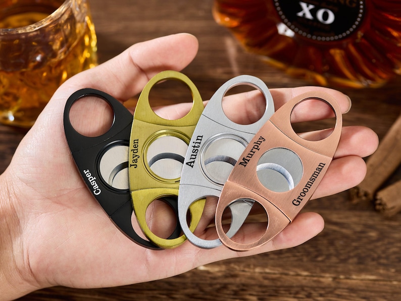 Pu&ograve; includere: Quattro taglia-sigari personalizzati nei colori nero, oro, argento e rame. Ogni taglia-sigari ha un foro circolare per il sigaro e un anello per il dito. I nomi sono incisi su ogni taglia-sigari.