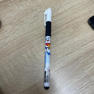 May include: A black and white pen with a white clip. The pen features a cartoon character design with blue polka dots and the text "MUC DEN 0.5mm". The pen is on a light brown wooden surface.