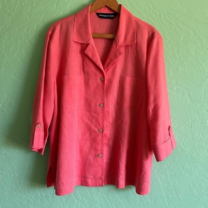 May include: A coral-colored button-down shirt with three-quarter sleeves, a collared neckline, and a chest pocket. The shirt is hanging on a wooden hanger against a light green wall.