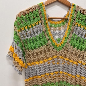 May include: A colorful crochet top with a V-neck design, featuring a mix of green, yellow, gray, and tan yarns. The open weave and short sleeves create a light and airy look. The top is displayed on a wooden hanger.