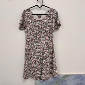 May include: A short-sleeved dress featuring a dense floral pattern with small pink, white, and black blossoms on a light background. The garment has a rounded neckline and a fitted bodice that flares slightly at the waist, hanging on a black hanger.