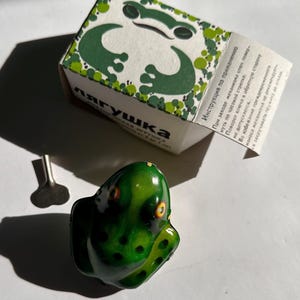 Könnte beinhalten: Ein Vintage-Aufziehspielzeug in Form eines Frosches, grün und schwarz lackiert, mit orangefarbenen Augen. Das Spielzeug wird von seiner Originalverpackung begleitet, die eine Froschillustration und Text in einer Fremdsprache aufweist. Ein kleiner Metallschlüssel ist ebenfalls enthalten.