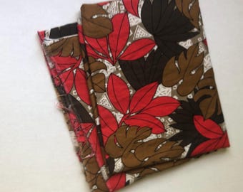 Puede incluir: Tela doblada con un estampado de hojas tropicales en rojo, marrón y negro sobre un fondo gris claro. La tela presenta un diseño repetitivo de hojas grandes y formas geométricas. Los bordes de la tela son visibles, mostrando los bordes crudos y sin terminar.