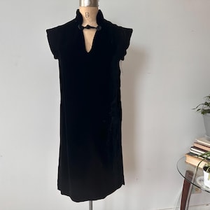 May include: A black velvet dress with a high collar and a V-neckline. The dress is sleeveless and falls to the mid-calf. The dress is displayed on a mannequin. The dress has a button closure at the neckline.