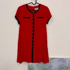 May include: A short-sleeved red dress featuring a black trim along the round neckline, sleeve cuffs, and a central vertical placket. The dress includes two faux chest pockets with black trim and a row of metallic buttons down the front center.