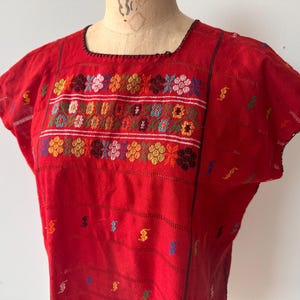 May include: A red, short-sleeved top with intricate floral embroidery in various colors across the chest. The top features a square neckline and horizontal stripes, showcasing a traditional textile design.