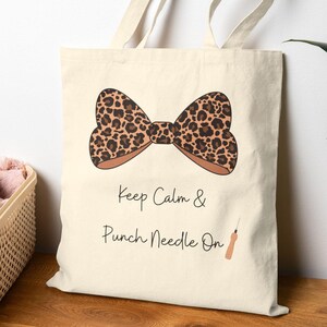 Może przedstawiać: Kremowa torba typu tote z wzorem kokardy w panterkę. Pod kokardą znajduje się napis "Keep Calm & Punch Needle On" oraz ilustracja narzędzia do punch needle.