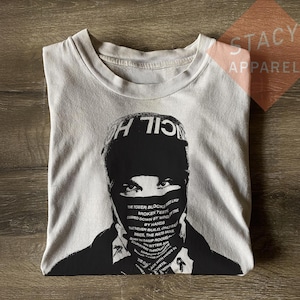 Könnte beinhalten: Hellgraues T-Shirt mit einer schwarz-weißen Grafik einer Person mit Maske und Hut. Auf dem Hut steht "CIL H". Die Maske und der untere Teil des Gesichts haben Text. Das T-Shirt ist auf einer Holzoberfläche gefaltet.