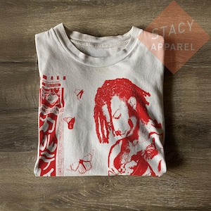 May include: White t-shirt with a red graphic print. The design features a portrait of a person with dreadlocks, butterflies, and text. The shirt is folded on a wooden surface. The text "STACY APPAREL" is visible.