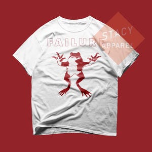 May include: White t-shirt with a red frog graphic and the word "FAILURE" printed across the top. The frog is in a stylized pose with its arms and legs spread out. The shirt is made of a soft, comfortable material.