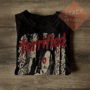 Puede incluir: Una camiseta negra de cuello redondo con un estampado gráfico que muestra la palabra terrified en letras rojas que gotean. El diseño incluye una ilustración en blanco y negro de un rostro con una marca circular roja sobre un ojo y flores.