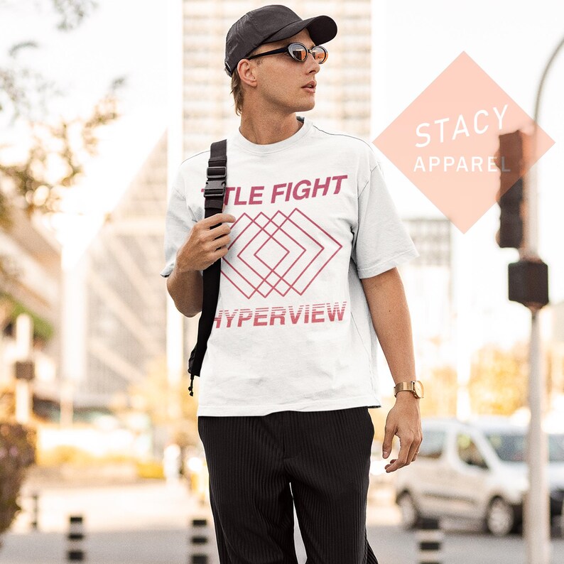 Puede incluir: Camiseta blanca con las palabras "TITLE FIGHT" y "HYPERVIEW" en fuente roja, junto con un dise&ntilde;o geom&eacute;trico. La persona lleva una gorra negra, gafas de sol y pantalones negros. El logotipo de Stacy Apparel es visible.