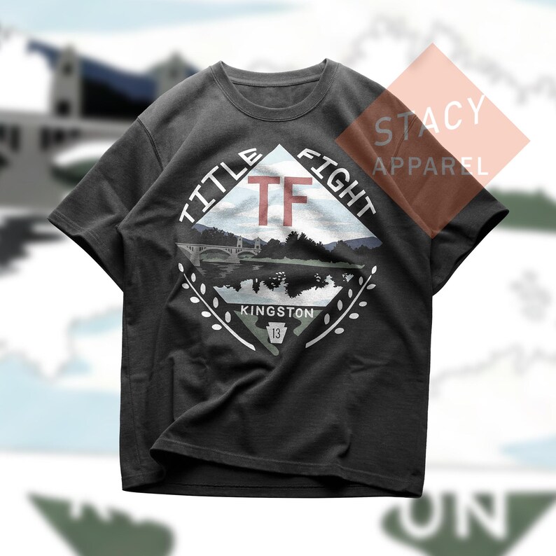 Puede incluir: Camiseta gris oscuro con un gr&aacute;fico en forma de diamante que muestra un puente sobre el agua, monta&ntilde;as y el texto "TITLE FIGHT" y "KINGSTON 13". La camiseta tambi&eacute;n tiene las iniciales "TF" en rojo.