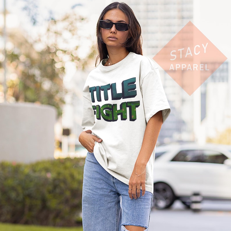 K&ouml;nnte beinhalten: Ein wei&szlig;es T-Shirt mit dem Schriftzug "TITLE FIGHT" in 3D-Gr&uuml;n und T&uuml;rkis. Das Shirt wird mit hellblauen Jeans kombiniert. Der Markenname "STACY APPAREL" ist oben rechts sichtbar.