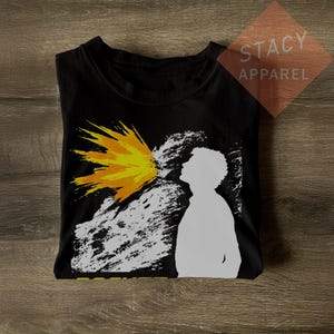May include: Black t-shirt with a graphic design featuring a white silhouette of a person exhaling a yellow and orange burst. The shirt has the text "STACY APPAREL" on a brown square.