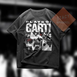 May include: Dark gray t-shirt featuring the text "PLAYBOI CARTI" in large white letters. The shirt has a graphic design with multiple images and symbols in white and red. The Stacy Apparel logo is visible in the upper right corner.