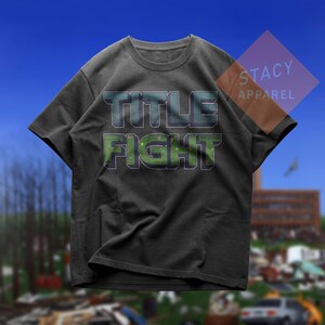 K&ouml;nnte beinhalten: Dunkelgraues T-Shirt mit dem Schriftzug "TITLE FIGHT" in Gr&uuml;n und Blau. Das Shirt hat einen Rundhalsausschnitt und kurze &Auml;rmel. Der Markenname "STACY APPAREL" ist oben rechts sichtbar.