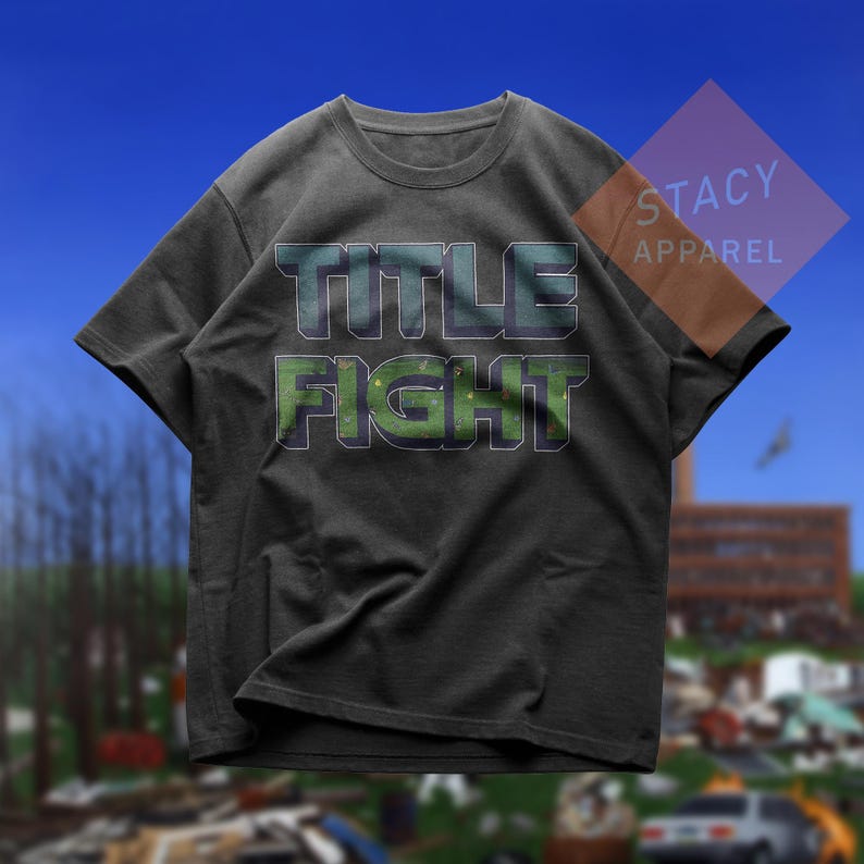 K&ouml;nnte beinhalten: Dunkelgraues T-Shirt mit dem Schriftzug "TITLE FIGHT" in Gr&uuml;n und Blau. Das Shirt hat einen Rundhalsausschnitt und kurze &Auml;rmel. Der Markenname "STACY APPAREL" ist oben rechts sichtbar.