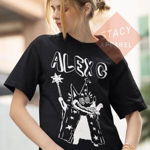 Puede incluir: Camiseta negra con el texto blanco "ALEX G" y un dibujo de un mago con una varita de estrella. El mago lleva un sombrero puntiagudo y una capa con patrones de estrellas. La camiseta es de Stacy Apparel.