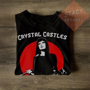 May include: Black t-shirt featuring the band name "CRYSTAL CASTLES" in white block letters above a red circle with a black and white graphic of a person. The shirt is from "STACY APPAREL".