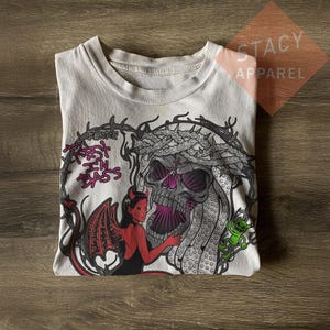 May include: A cream-coloured t-shirt featuring a graphic print of a large skull wearing a crown of thorns, a red devil figure with wings, and a small green creature. The text "Encore" and "Rest In Bass" is printed in pink and black lettering.
