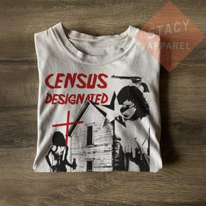 Puede incluir: Camiseta blanca con un diseño gráfico que presenta el texto "CENSUS DESIGNATED" en rojo, una casa, una cruz y una mujer con una pistola. La camiseta está doblada sobre una superficie de madera.