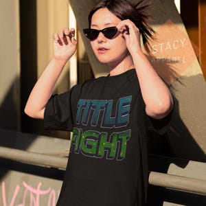 K&ouml;nnte beinhalten: Schwarzes T-Shirt mit den Worten "TITLE FIGHT" in einem 3D-Design. Der obere Text ist t&uuml;rkis und blau, und der untere Text ist gr&uuml;n. Das T-Shirt wird von einer Person getragen, die eine schwarze Sonnenbrille tr&auml;gt.
