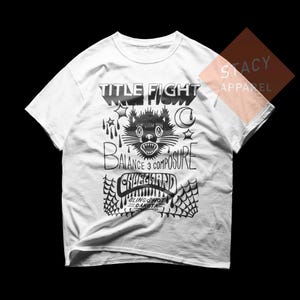 May include: White t-shirt with a black graphic design. The design features the words "TITLE FIGHT" above a cat illustration, with the words "BALANCE & COMPOSURE" and "CHUCKLERD" below. The text "SLINGSHOT DAKOTA" is also included.