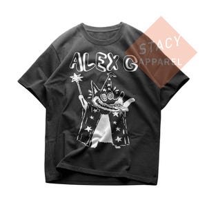 Op de afbeelding: Donkergrijs t-shirt met een witte cartoon toverhond met een toverstaf en een tovenaars hoed. De tekst "ALEX G" staat boven de afbeelding. Het shirt is van Stacy Apparel.