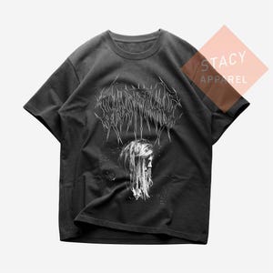 May include: Dark gray t-shirt featuring a white graphic design. The design includes stylized text above a detailed illustration of a person with long hair. The shirt has short sleeves and a crew neck. The brand name "STACY APPAREL" is visible.