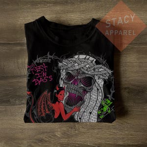 Peut inclure: Un t-shirt noir avec une impression graphique représentant un crâne portant une couronne d'épines, un personnage diable rouge et une petite créature verte. Le texte indique Encore et Rest In Bass en rose, entouré de barbelés et d'éléments sombres.