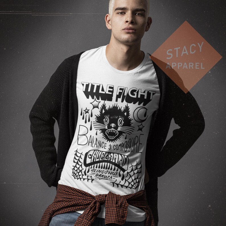 May include: White t-shirt with a black graphic design featuring a cat, stars, and the words "TITLE FIGHT", "BALANCE 3 COMPOSURE", and "CHUEGARD SLINGSHOT DAKOTA". The shirt is worn with a black cardigan and a red and brown plaid shirt tied around the waist.