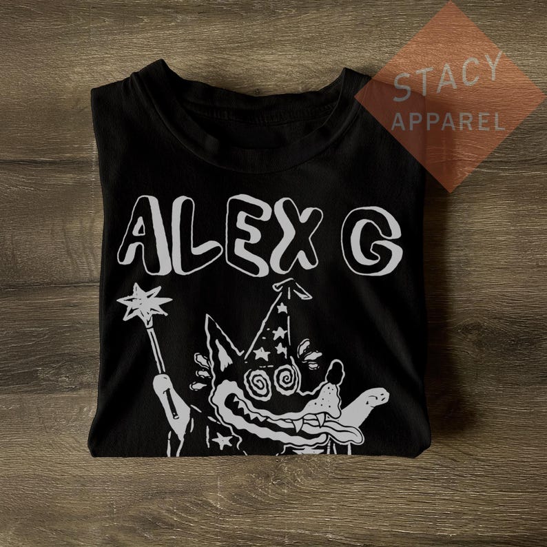 Puede incluir: Camiseta negra con el texto blanco "ALEX G" sobre un lobo de dibujos animados con un sombrero de mago y una varita. La camiseta es de Stacy Apparel.