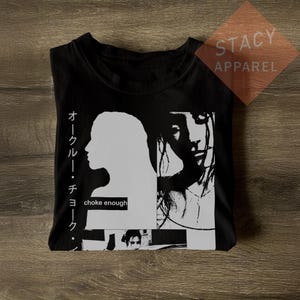 Puede incluir: Camiseta negra con un diseño gráfico que presenta una silueta blanca de un perfil y una cara, junto con el texto "choke enough" y caracteres japoneses. La camiseta también tiene el texto "STACY APPAREL".