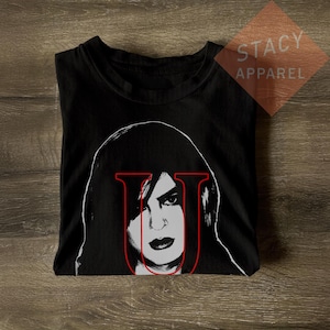 May include: A black t-shirt featuring a high-contrast white graphic of a person with dark hair, overlaid with a bold red letter U. The design is centered on the chest, showcasing a stylized portrait with sharp lines and a minimalist color palette.